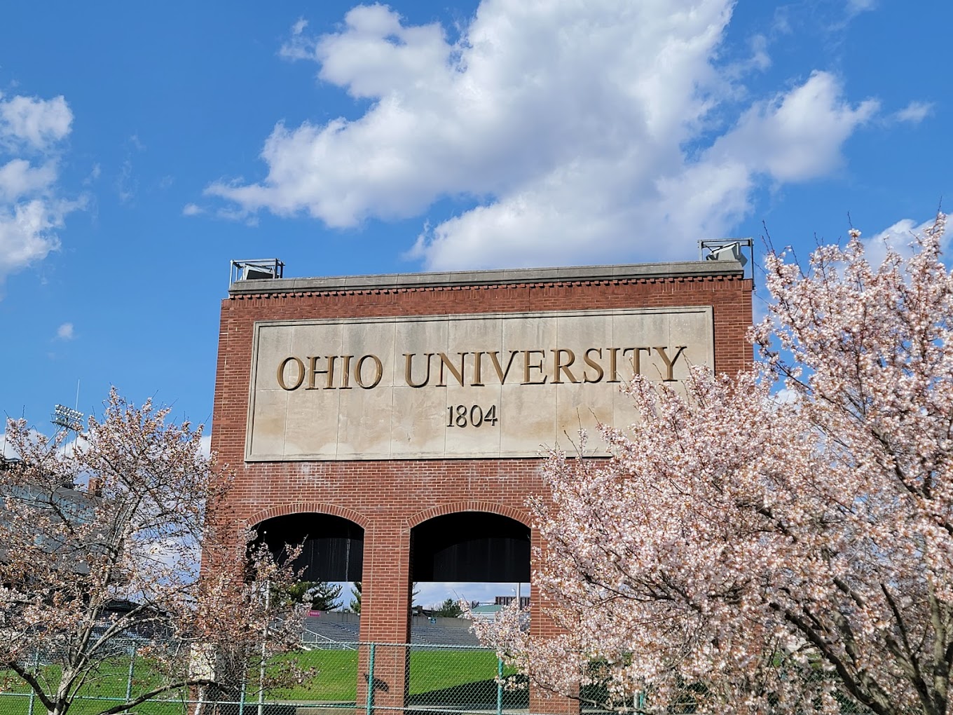 Brick archway with "Ohio University 1804" and blooming trees beneath a blue sky.